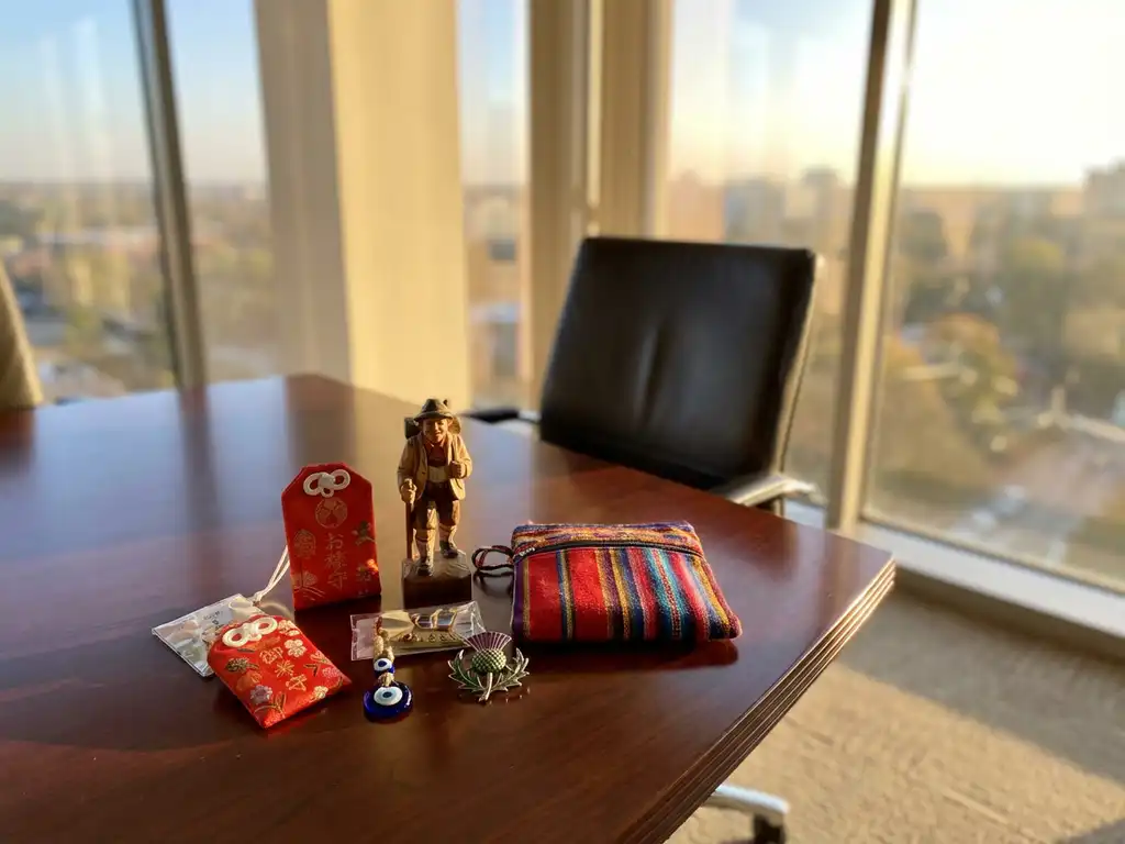 Abschiedsgeschenke aus verschiedenen Ländern auf Konferenztisch - japanischer Omamori-Glücksbringer, deutsche Holzfigur und buntes lateinamerikanisches Textil bei warmem Sonnenlicht