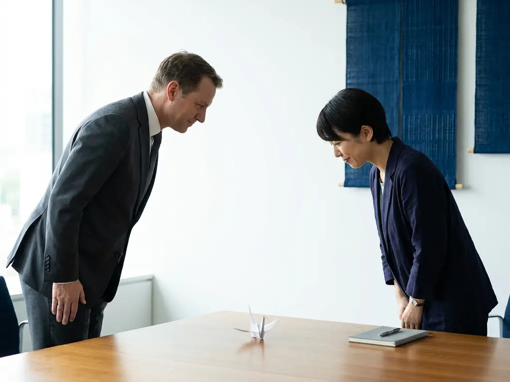 Deutscher und japanischer Geschäftspartner verbeugen sich zur Begrüßung am Konferenztisch mit Origami-Kranich und Notizbuch.